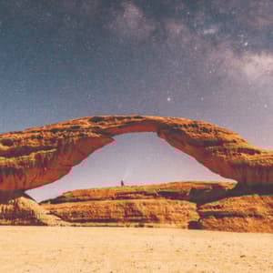 Una persona solitaria si erge sotto un imponente arco roccioso naturale nel deserto, con la Via Lattea visibile nel cielo notturno stellato.
