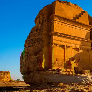 Una tomba monumentale scavata nella roccia arenaria risplende sotto il sole dorato in un vasto deserto sotto un cielo azzurro e limpido.