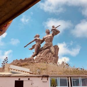 Une statue monumentale en bronze représentant une famille se dresse au sommet d'une colline rocheuse sous un ciel bleu parsemé de nuages blancs et cotonneux.