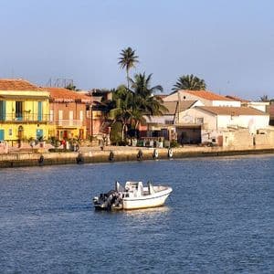 Un bateau à moteur blanc sur l'eau, devant des bâtiments colorés en bord de mer et des palmiers sous un ciel dégagé.
