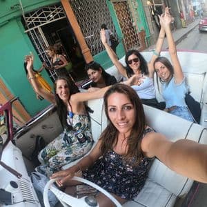 Eine WeRoad Frauengruppe macht ein lachendes Selfie in einem pinkfarbenen Oldtimer-Cabrio auf der Straße.