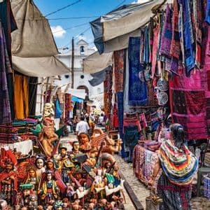 Un marché de rue animé, débordant de textiles colorés et à motifs et d'artisanat en bois sculpté, sous un ciel ensoleillé.