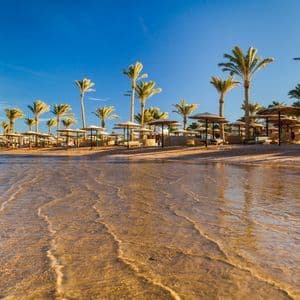 Flaches, klares Wasser umspült einen Sandstrand mit Palmen und Strohschirmen unter blauem Himmel.