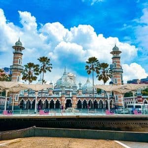 Una moschea sontuosa con minareti e cupole a strisce si erge oltre un fiume, con palme e grandi ombrelloni bianchi in primo piano.