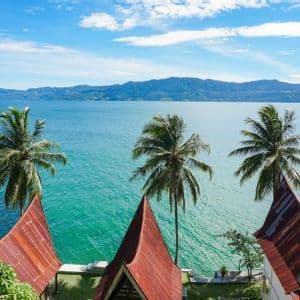 Tetti rossi appuntiti e palme si affacciano su una vasta distesa di acqua turchese con montagne sullo sfondo sotto un cielo azzurro.