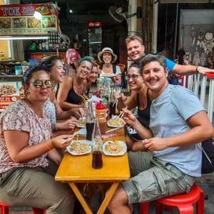Un groupe WeRoad sourit en dégustant des nouilles avec des baguettes à un stand de street food en plein air.
