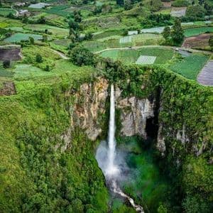 Vista aerea di un'imponente cascata che si getta in una gola verdeggiante, circondata da un mosaico di campi coltivati verdi.