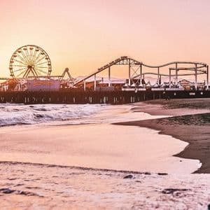 An amusement park pier with a Ferris wheel and roller coaster on a beach, viewed as waves wash ashore during sunset.