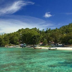 Una vista di una baia tropicale con acqua turchese cristallina, una spiaggia di sabbia bianca con barche e un villaggio immerso in una collina boscosa.