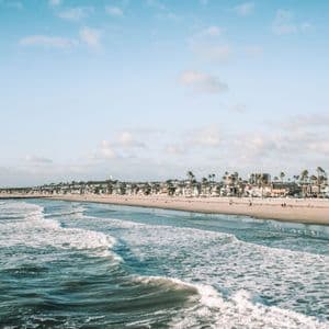 Le onde dell'oceano si infrangono su un'ampia spiaggia sabbiosa costeggiata da palme ed edifici sotto un cielo azzurro con nuvole leggere.