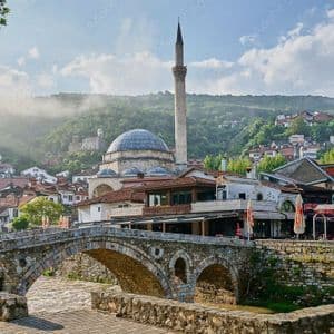 A stone arch bridge spans a river before a town with a mosque and minaret, with a misty green hill in the background.