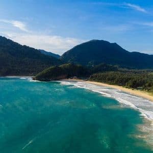 Vista aerea di una spiaggia sabbiosa e ricurva con onde turchesi, circondata da lussureggianti montagne verdi e foreste sotto un cielo azzurro.