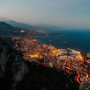 Veduta dall'alto di una città costiera illuminata e del suo porto turistico al tramonto, incastonata tra mare e monti.