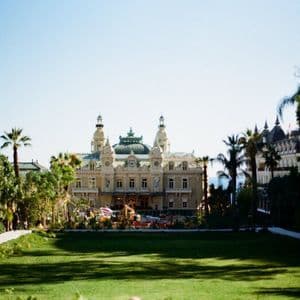 Una vista del Casinò di Monte Carlo da un parco, con un grande prato verde in primo piano e palme che incorniciano l'edificio.