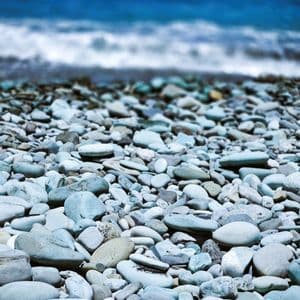 Nahaufnahme von glatten, hellblauen Kieselsteinen am Strand, mit dem tiefblauen Meer unscharf im Hintergrund.