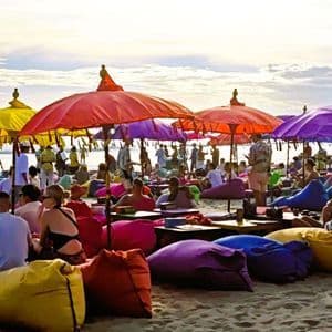 Eine große Menschenmenge entspannt sich auf farbigen Sitzsäcken unter bunten Sonnenschirmen an einem Sandstrand.