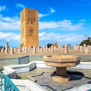 A tall, square, sand-colored stone tower stands behind rows of ancient columns in a plaza, with an ornate fountain in the foreground.