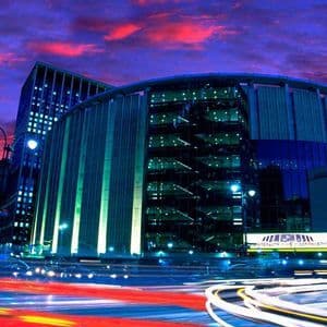 Lunga esposizione di una strada cittadina di notte, con scie luminose del traffico davanti a moderni edifici illuminati sotto un cielo viola.