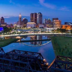 Uno skyline cittadino illuminato e un moderno ponte bianco riflessi in un fiume al crepuscolo, con un ponte ferroviario in primo piano.
