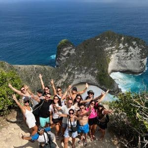 Un groupe WeRoad en voyage sourit et pose sur un point de vue de falaise avec une formation rocheuse spectaculaire et l'océan turquoise en contrebas.