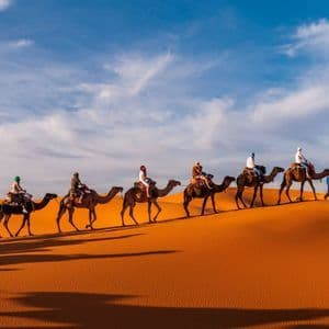 A WeRoad group trip rides a line of camels over orange desert sand dunes under a partly cloudy sky.
