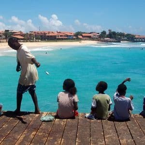 Un ragazzo su un pontile di legno accanto a tre bambini seduti, con un piccolo pesce sospeso sull'oceano turchese.