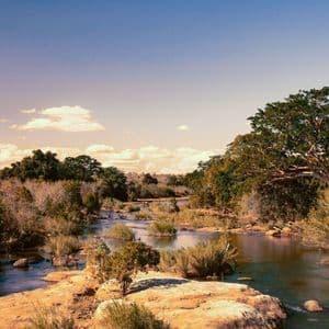 Un ampio fiume scorre attraverso un paesaggio roccioso circondato da fitti cespugli e alberi sotto un cielo azzurro limpido con nuvole sparse.
