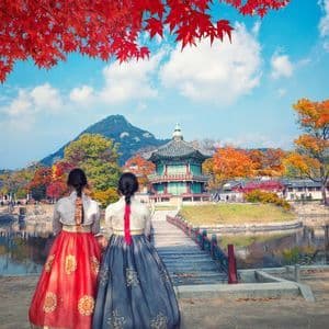 Due donne in abiti tradizionali hanbok guardano un lago verso una pagoda circondata da un vibrante fogliame autunnale.