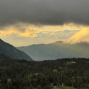 Ein üppiges grünes Tal und bewaldete Berge unter einem dunklen, bewölkten Himmel mit durchbrechendem goldenem Sonnenlicht.