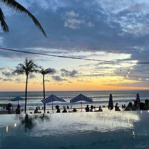 Eine WeRoad-Gruppe beobachtet den Sonnenuntergang von einem Infinity-Pool aus mit Blick auf einen Strand mit Palmen und Lichterketten.