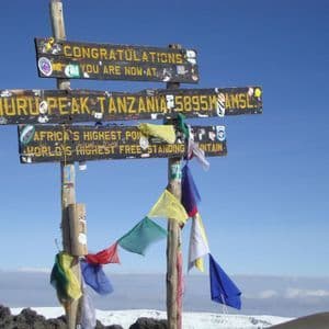 The wooden sign at Uhuru Peak, Tanzania, marking it as Africa's highest point, stands against a clear blue sky above the clouds.