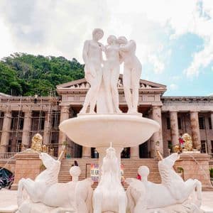 Una gran fuente con estatuas clásicas blancas se alza frente a un edificio columnado con andamios, teniendo como telón de fondo una colina boscosa.