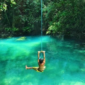 Una persona vista de espaldas en un columpio de cuerda se desliza sobre agua turquesa, con un denso bosque verde de fondo.