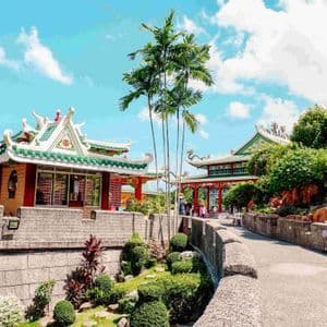 Un complejo de templos asiáticos tradicionales con techos de tejas verdes está rodeado de palmeras y jardines cuidados bajo un cielo azul.