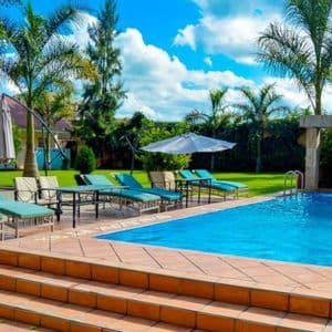 A swimming pool with a waterfall feature, surrounded by empty lounge chairs, palm trees, and a green lawn under a blue sky with clouds.
