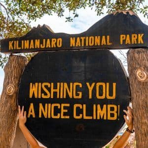 A person's hands hold the wooden sign for Kilimanjaro National Park, which reads, "Wishing you a nice climb!".