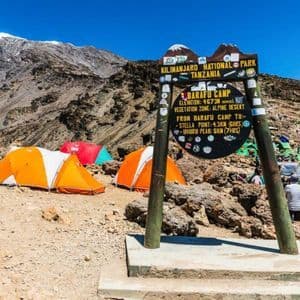 A wooden signpost stands at a mountain base camp, with colorful tents pitched on rocky terrain and a snow-capped peak in the background.