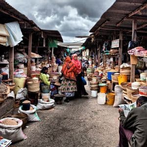 A busy outdoor market alley with people browsing stalls filled with grains and spices in woven baskets.