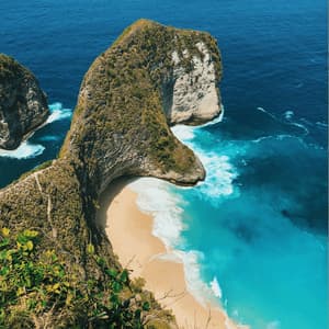 A high-angle view of a large, green-covered cliff formation curving over a secluded sandy beach and turquoise ocean.