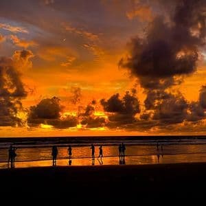 Silhouettes d'un voyage de groupe WeRoad sur une plage, observant un coucher de soleil orange dramatique sur l'océan avec des nuages sombres.