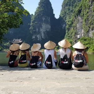 Un grupo de viaje WeRoad, sentado en una saliente de piedra, con sombreros cónicos, contempla un río rodeado de exuberantes montañas verdes.