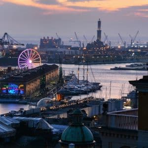 Una vista aerea di un porto al crepuscolo, che mostra una ruota panoramica illuminata, un faro, barche ormeggiate e gru industriali.
