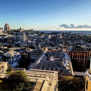 Una vista elevata diurna sui tetti di una fitta città costiera, con un porto e il mare visibili in lontananza.