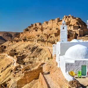 Una moschea imbiancata a calce con una cupola e un minareto si erge di fronte a un villaggio costruito su un fianco di collina desertica, sotto un cielo azzurro.