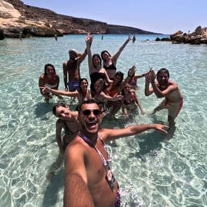 Un gruppo WeRoad sorride e posa per un selfie di gruppo, immerso nelle acque limpide e poco profonde di una costa rocciosa.