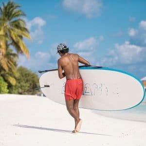 Un hombre con pantalones cortos rojos y gorra lleva una tabla de paddle surf por una playa de arena blanca con palmeras.