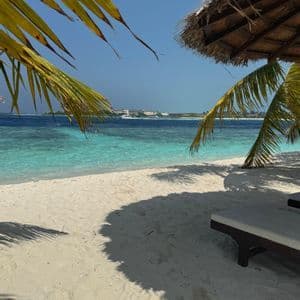Una tumbona y una sombrilla de paja en una playa de arena blanca, enmarcada por hojas de palma, con vistas a un océano turquesa.