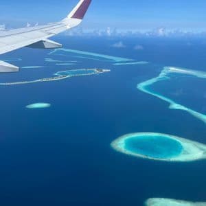 Una vista aérea desde la ventanilla de un avión, mostrando el ala sobre un océano azul profundo con varios atolones turquesa y bancos de arena.