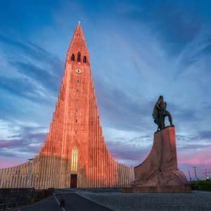 Eine hohe, moderne Kirche und eine Statue auf einem Sockel werden vom warmen Schein des Sonnenuntergangs vor einem dramatischen, bewölkten Himmel beleuchtet.