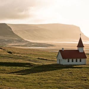 Eine weiße Kirche mit rotem Dach und Turm steht auf einem grasbewachsenen Feld mit dunstigen Klippen und einer Küste im Hintergrund.